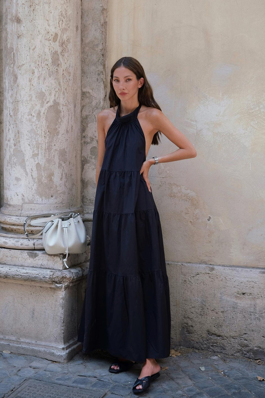 A woman standing in front of an old building wearing a black linen halter maxi dress with black slides and a white bucket bag.