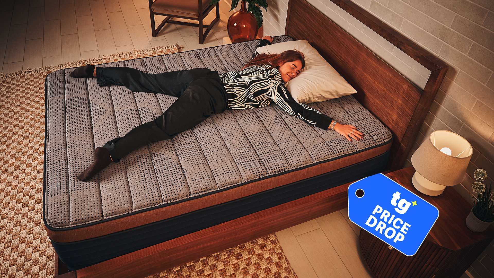 Mattress tester Eve Davies stretches out on the Brooklyn Bedding CopperFlex Memory Foam Mattress on a bed frame in a warmly lit room. In the bottom right corner, a Tom's Guide price drop badge