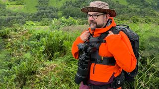 Photographer Dan M Lee wearing a red coat and zoom lens with a grassy background