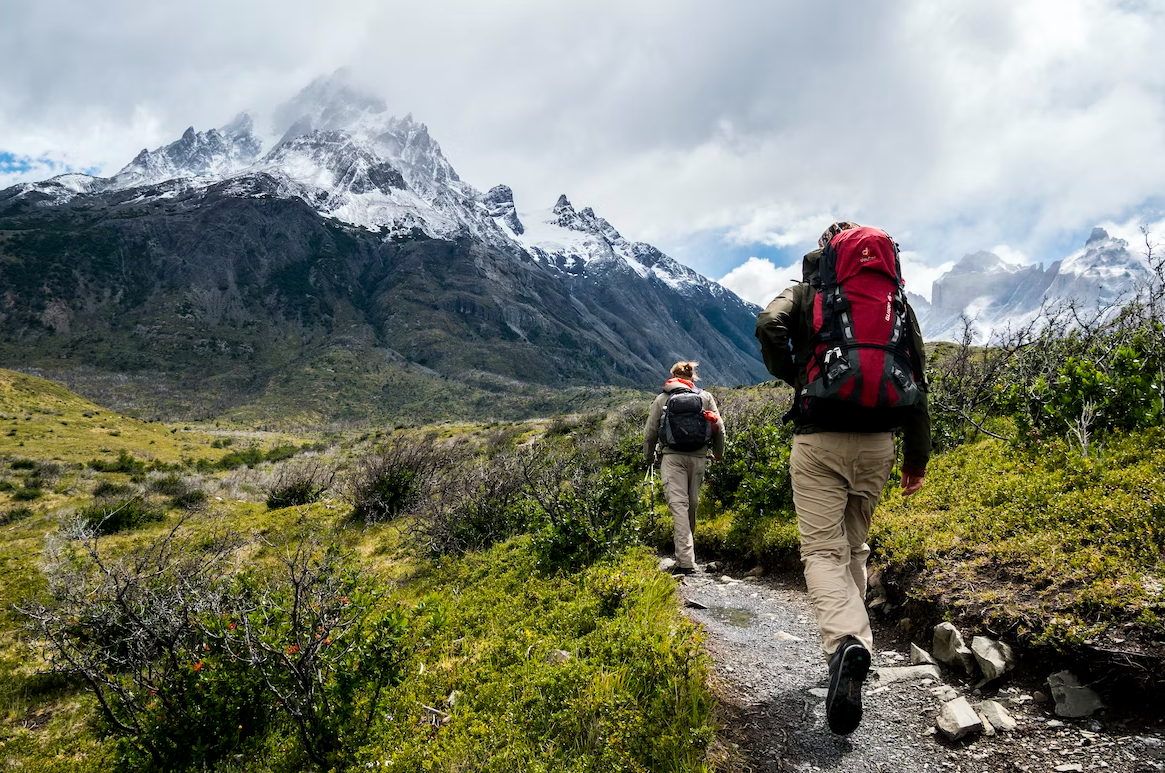 A pair of hikers are moving towards the mountains, they carry packs and are dressed for cooler weather in pants and jackets. 