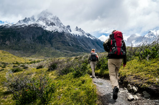 A pair of hikers are moving towards the mountains, they carry packs and are dressed for cooler weather in pants and jackets.