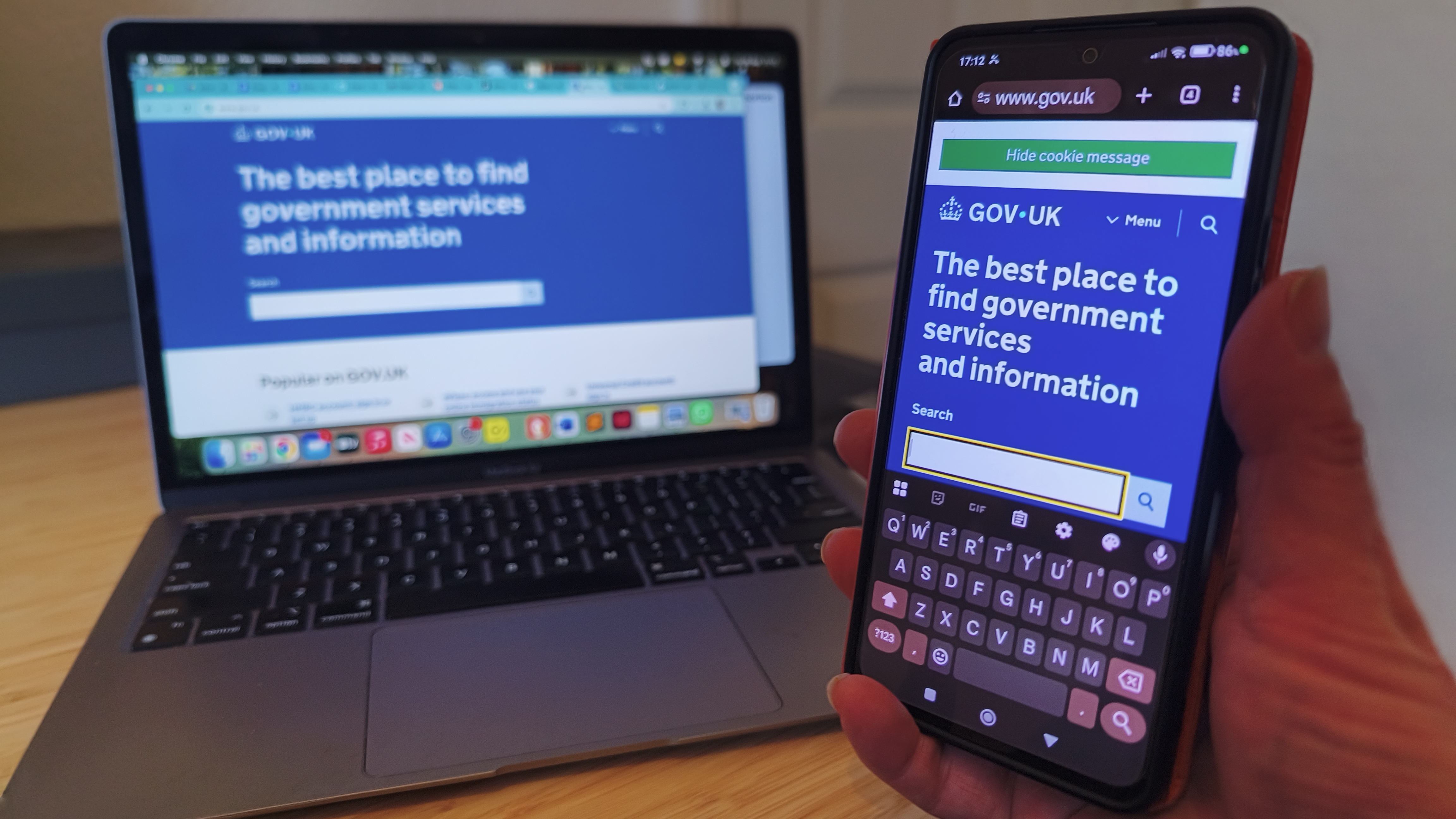 A hand holds a smartphone displaying the GOV.UK website, which is also open on a laptop in the background, both showing the text &quot;The best place to find government services and information.&quot;