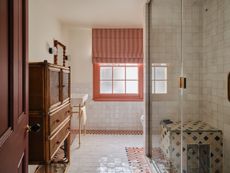 Bathroom with cream wall and floor tiles, a colorful striped tile border, mosaic shower seat, vintage cane cabinet, and coral-painted window frame.