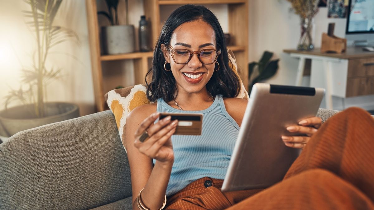 A young woman is smiling while sitting on a lounge using a tablet. She&#039;s holding a credit card in her hand.