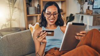 A young woman is smiling while sitting on a lounge using a tablet. She's holding a credit card in her hand.