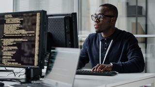 Male software engineer working at a computer in open plan office space with PC screen with code pictured in foreground.