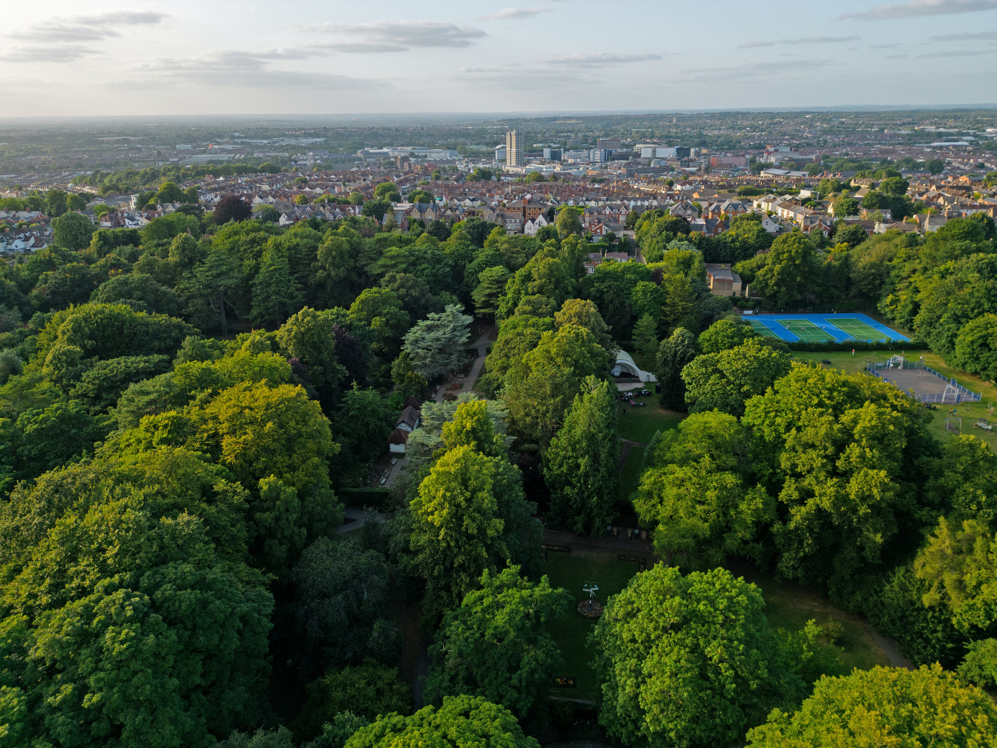 Panoramic view of Swindon town center. Aerial above botanical tree canopy at sunset. View from Swindon Town Gardens public park with recreational grounds and tennis court.