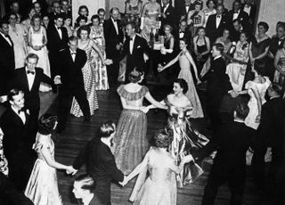 A black and white photo of Lady Pamela Mountbatten and Queen Elizabeth II dancing in the middle of people wearing tuxedos and gowns