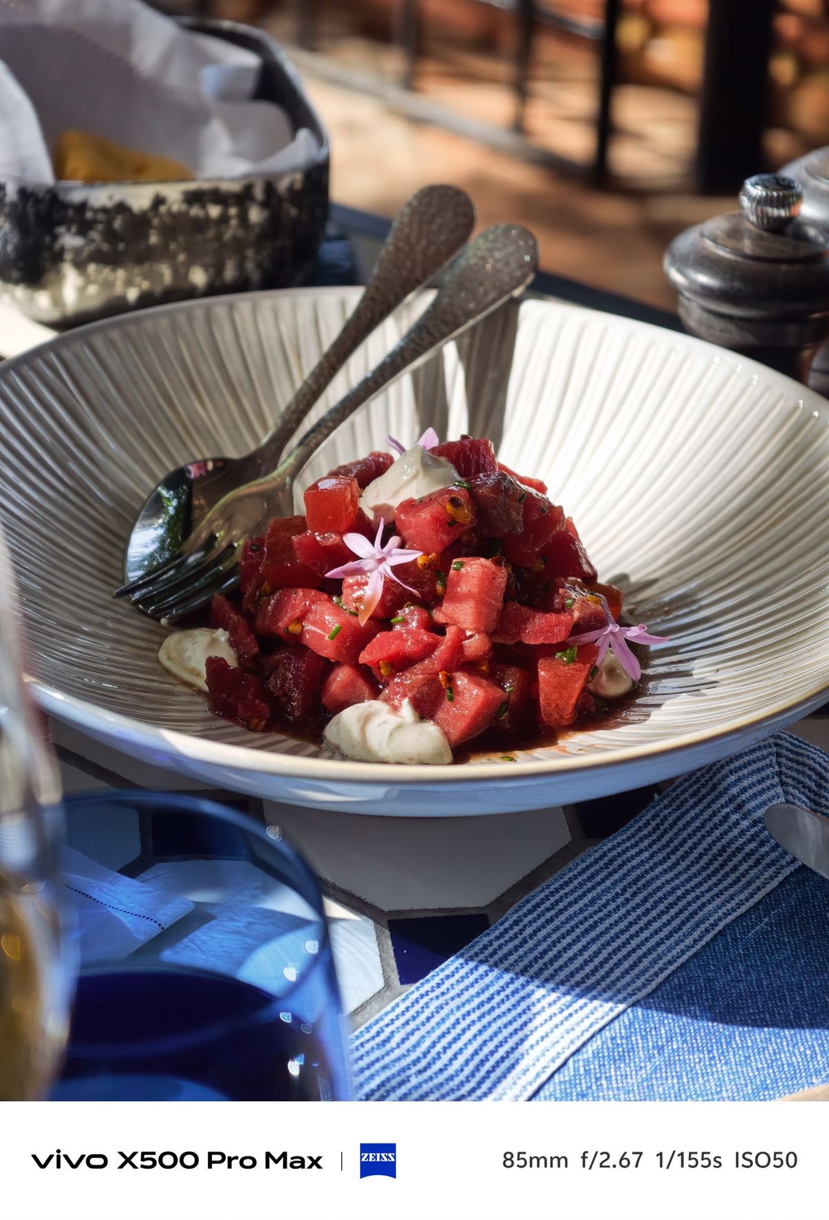 Bowl of finely diced red meat garnished with edible flowers and creamy sauce on an outdoor restaurant table.