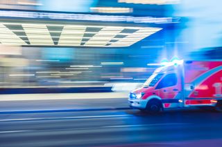 An ambulance going at high speed with its flashing lights turned on. The background behind it is blurred