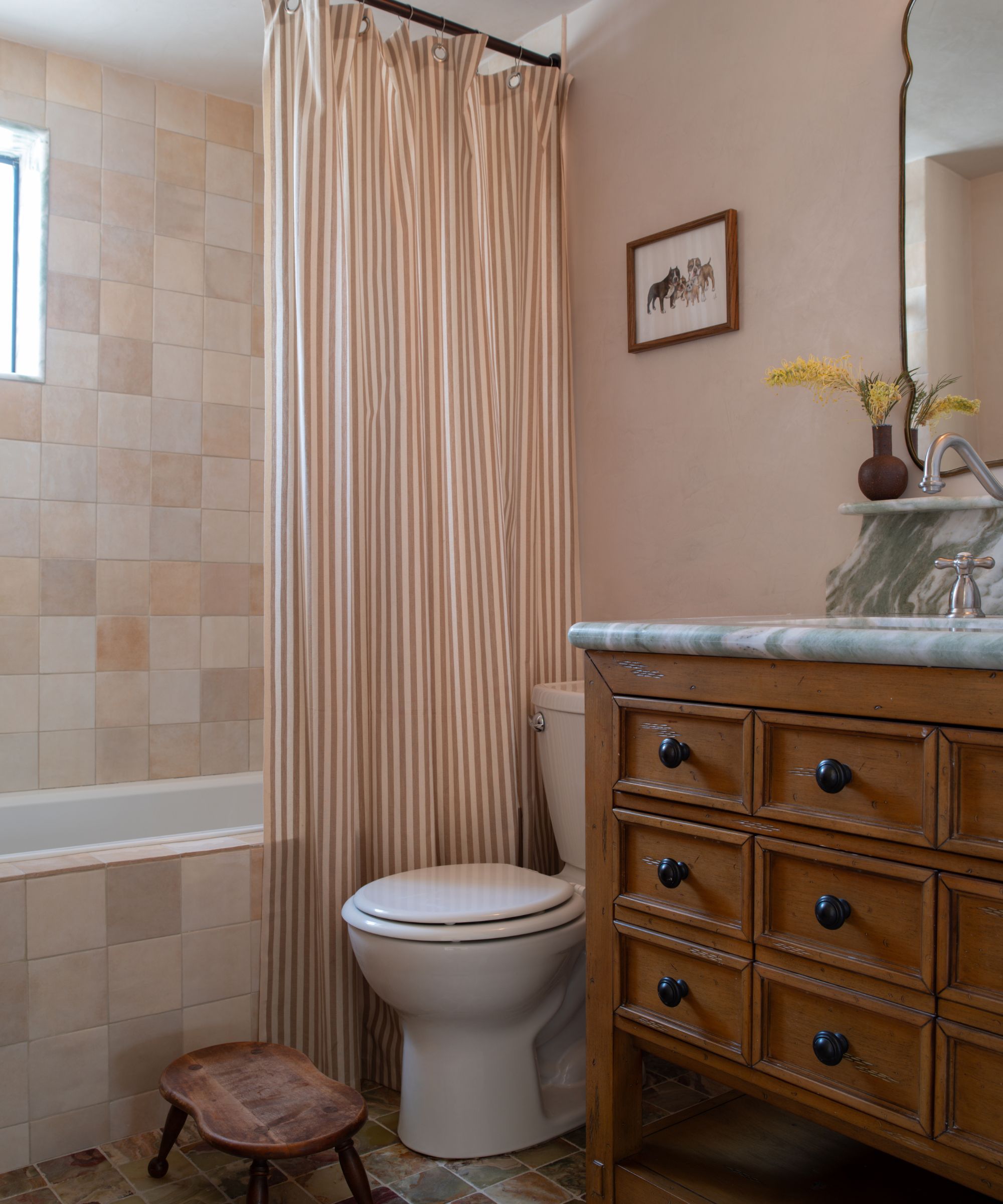 small bathroom with pale pink walls, tiles, and striped shower curtain, wooden vanity unit, and marble counter