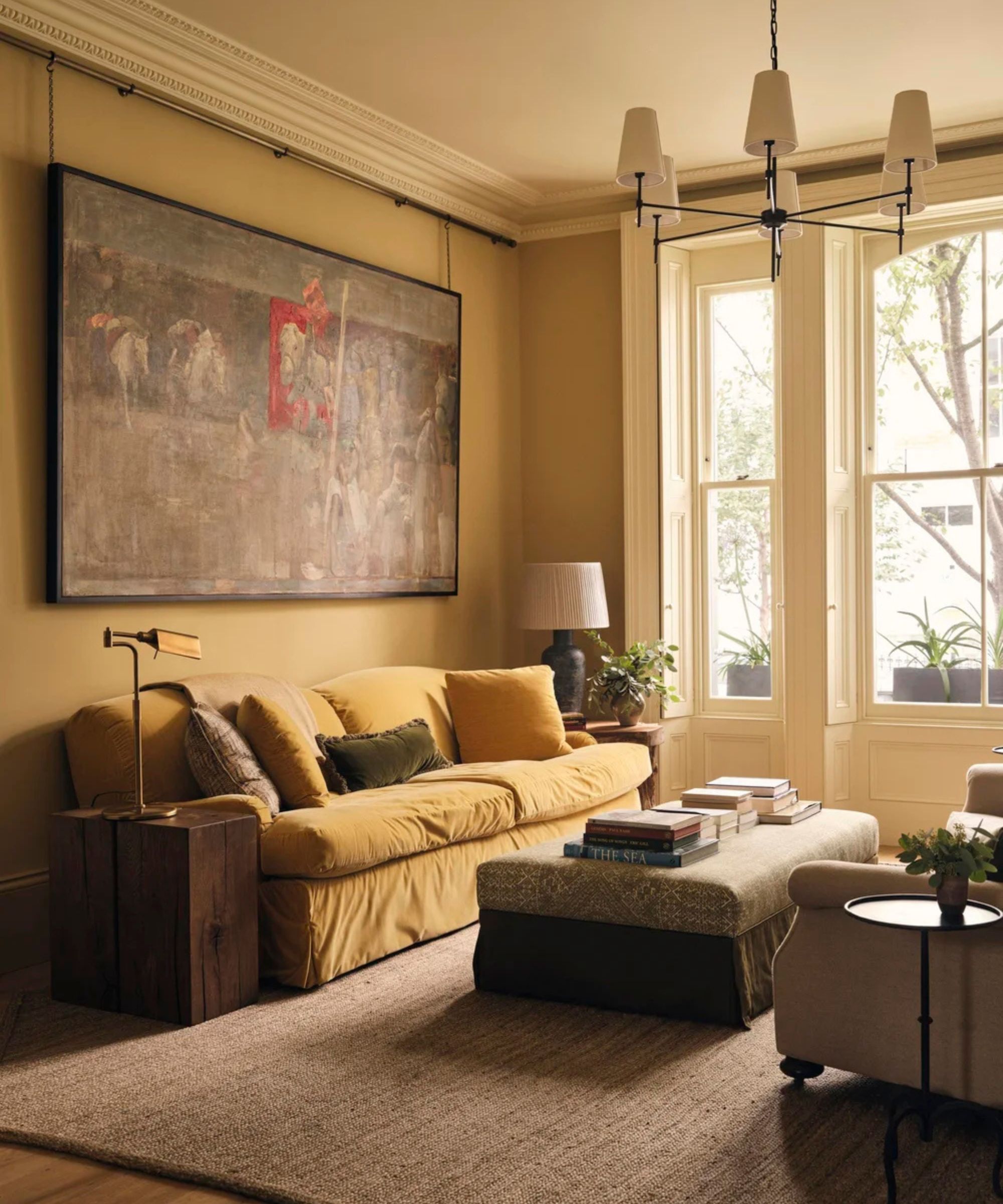 A warm-toned living room with ochre walls features a yellow velvet sofa, a large vintage painting hanging on a rail, and an ottoman coffee table in front of tall bay windows