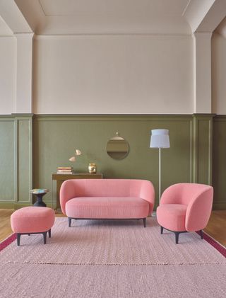 living room with green accent wall, cream walls, pink modern couch and armchair, footstool, lamp, rug, console, mirror