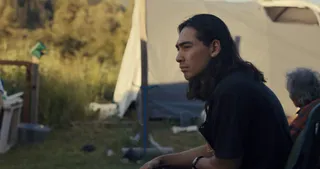 A man with long hair sits outdoors in front of a tent and a wooden stake, with an older man behind him (right), in 'Sugarcane.'