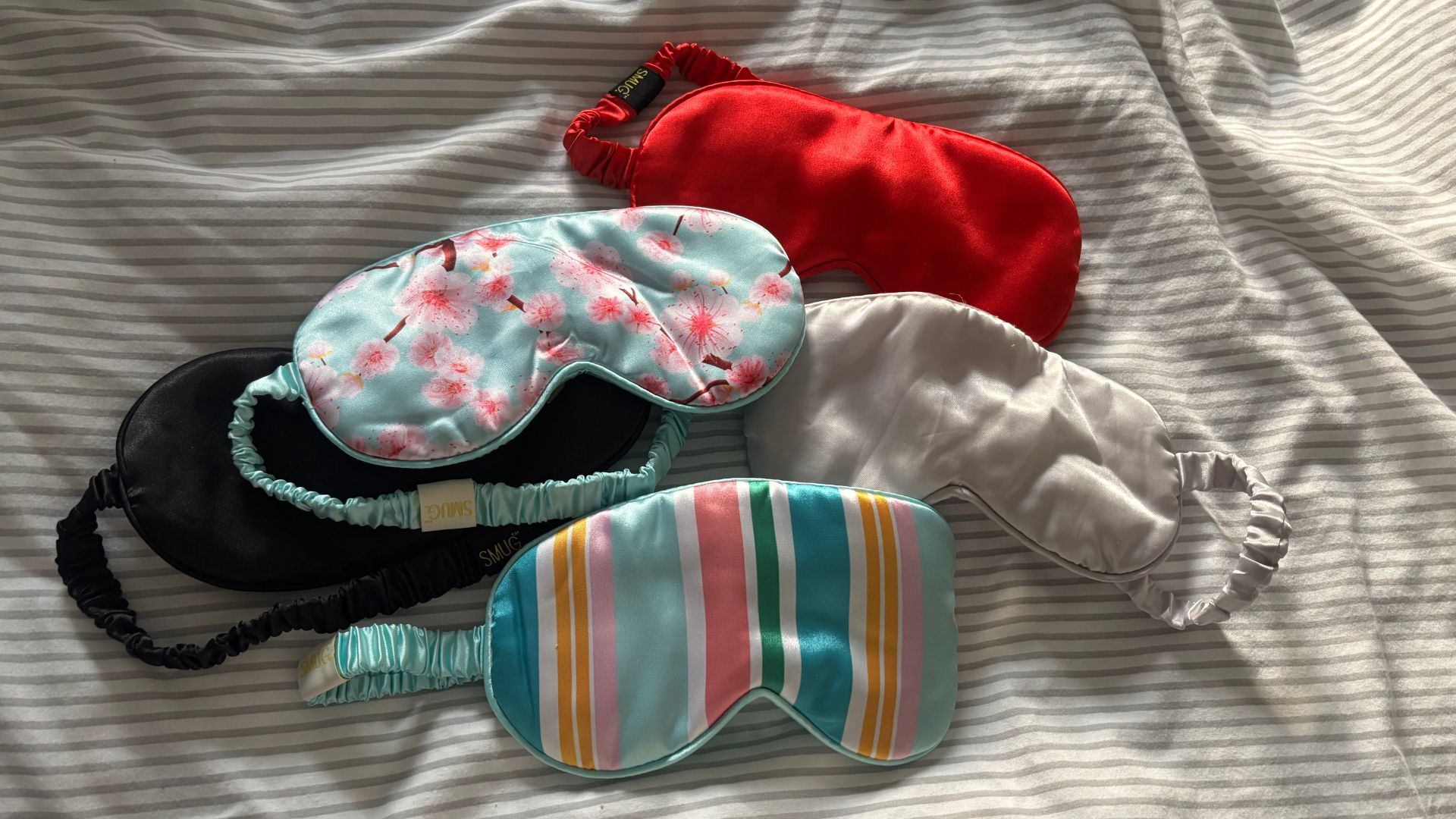 A collection of SMUG Satin Sleep Masks in a range of patterns and colours spread out on striped bedding.