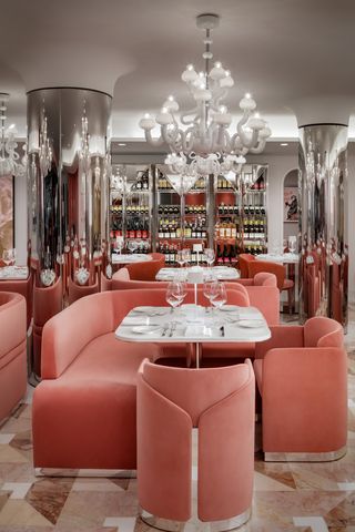 A lavish dining room features marble tables surrounded by soft pink, velvety banquettes, and Murano glass chandeliers in pale white.