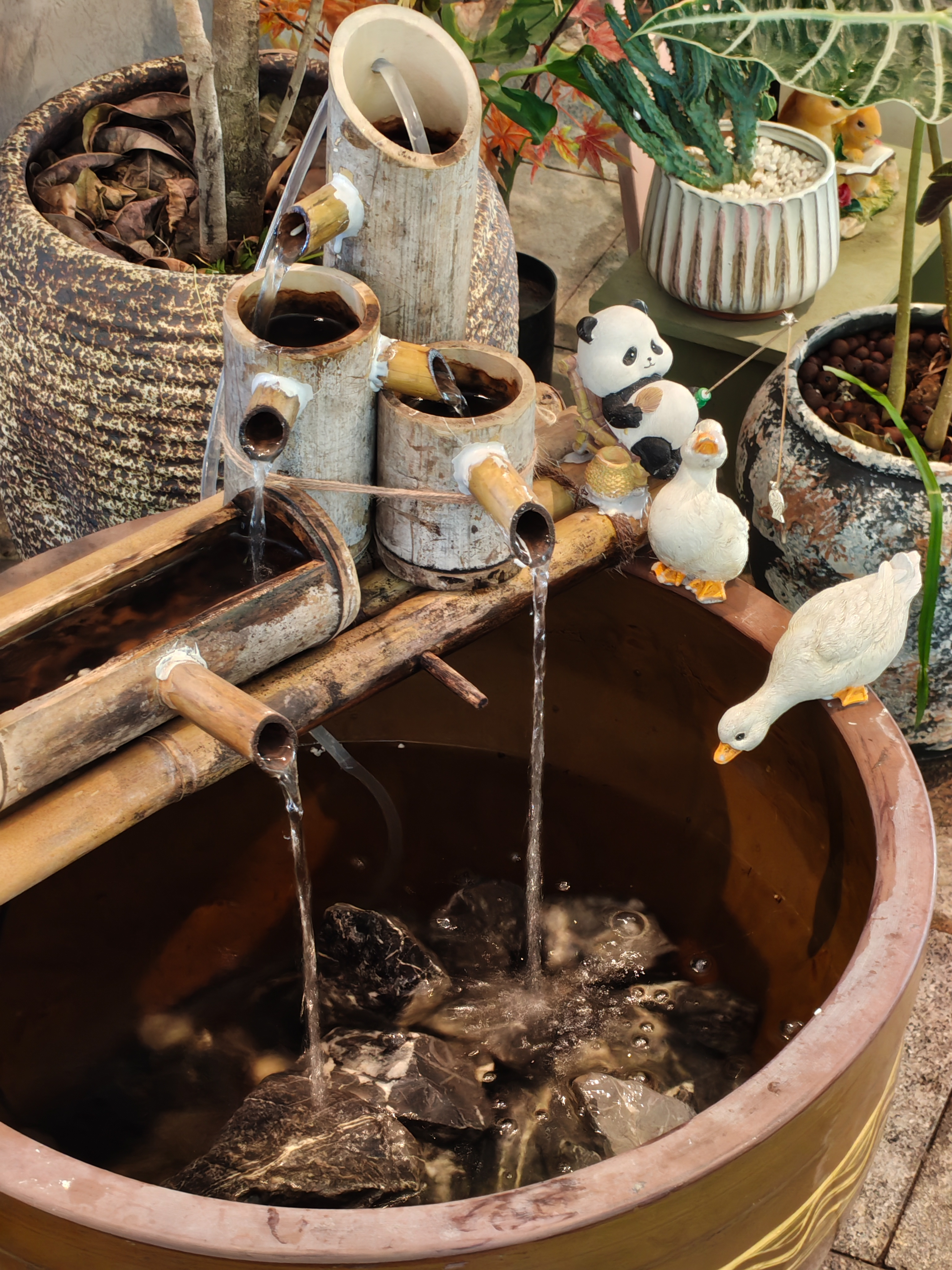 A decorative indoor bamboo water fountain. Water flows from several tiered bamboo pipes into a large, dark ceramic basin filled with river rocks. Small ceramic figurines, including a panda and two white ducks, are perched on the bamboo and the edge of the basin. Other potted plants are visible in the blurred background.