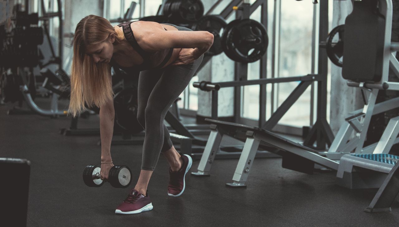 Woman performing B-stance Romanian deadlift in gym. She is holding one dumbbell, one foot is in front of the other and she is bent over with a straight back.