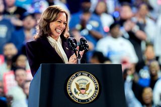 Kamala Harris stands at the podium at a rally wearing a purple blazer with a bow tied blouse