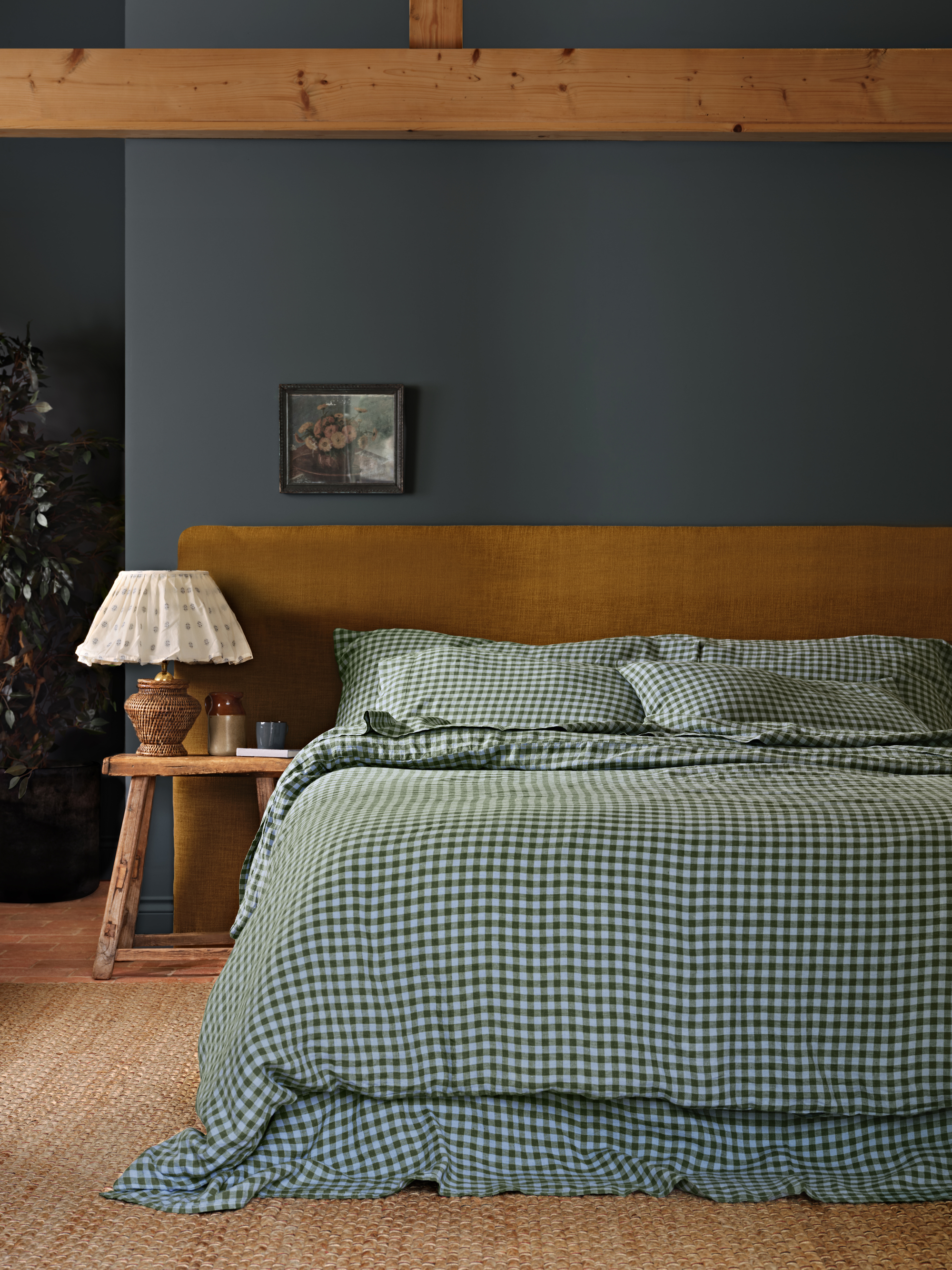 Image of a bed with an orange headboard and a blue and green gingham bedding set. The walls of the room are dark blue and there is a rattan rug.