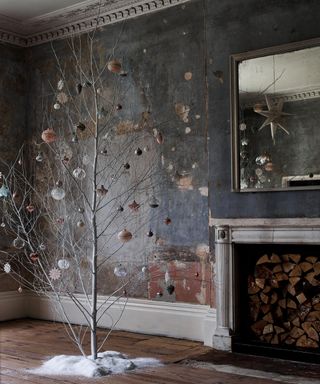 A twig tree decorated with baubles in industrial rustic room with peeled walls, wooden floorboards and unused fireplace
