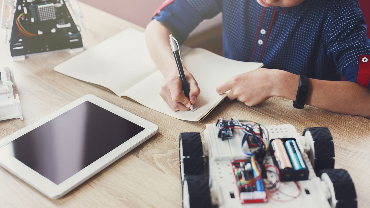 A child working on a robotics project sitting at a desk with a tablet and two robots writing on a notepad