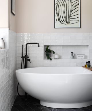 white oval shaped bath in corner of bathroom with white tiles, pink painted walls above and black floor, black floor mounted bath tap