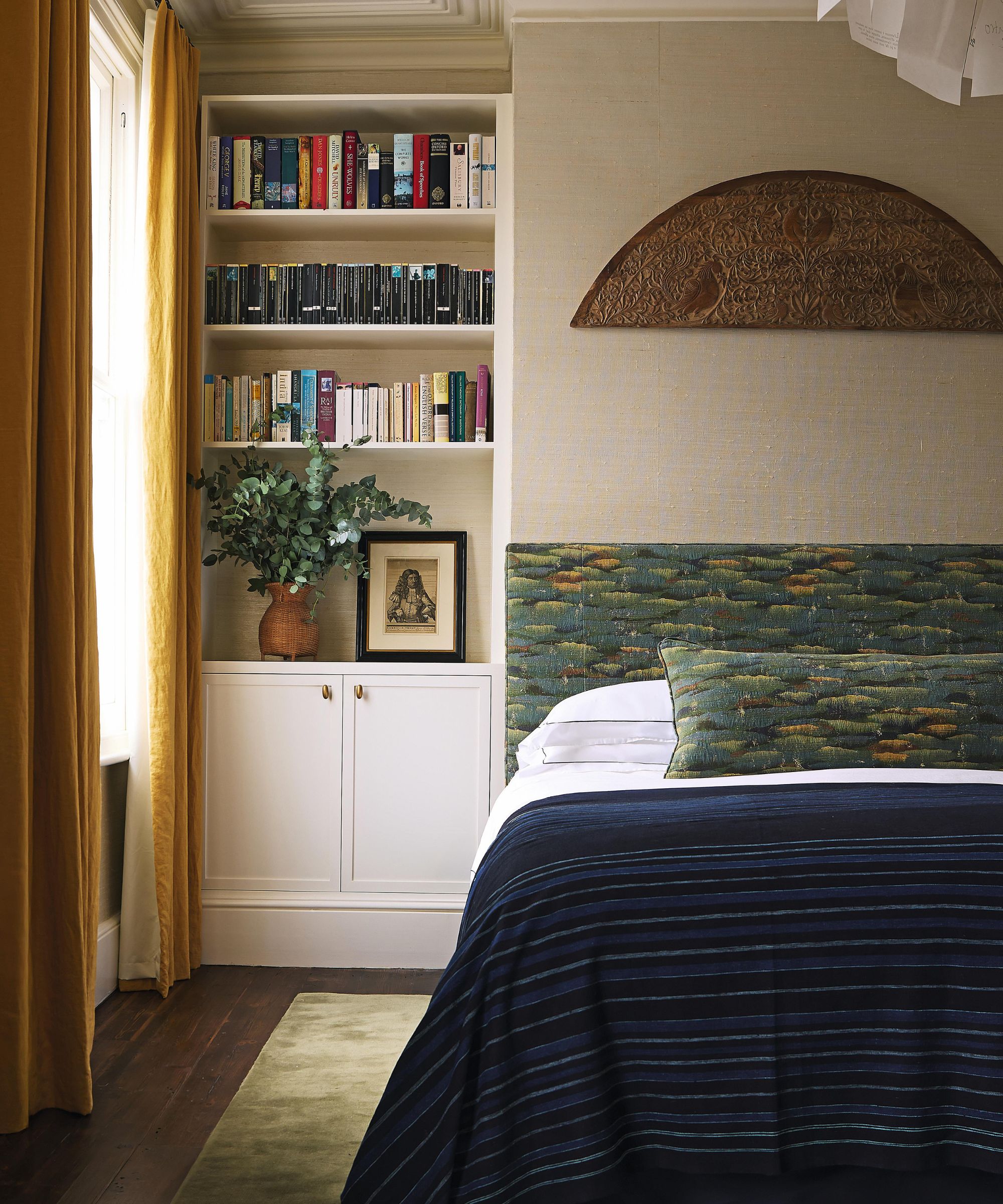 a neutral london bedroom with a bed place on the chimney breast, alcove bookcase and cupboard, yellow drapes at the window, and a green patterned custom upholstered headboarrd