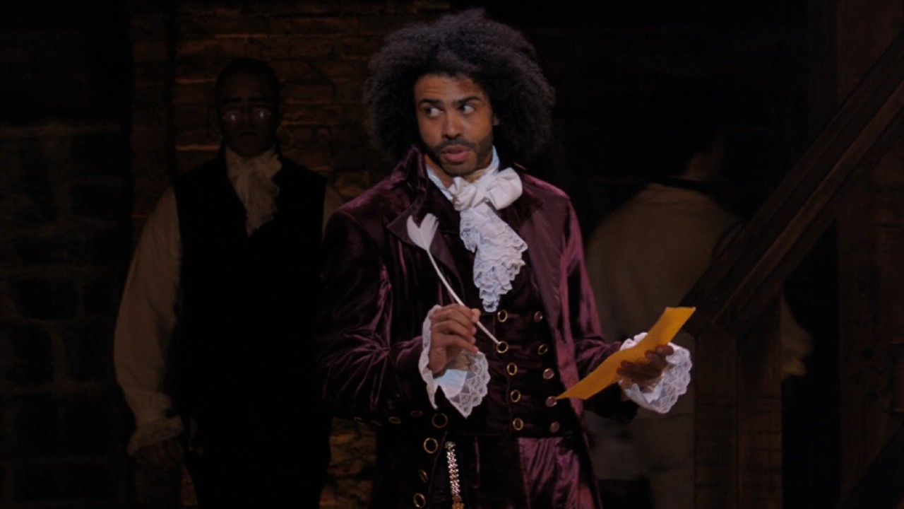 Daveed Diggs as Jefferson holding a quill in his left hand, parchment in the right. He's giving the side eye to the left.