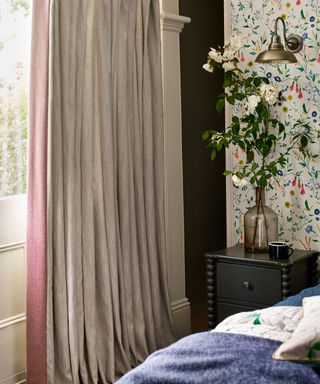 Close up of window in bedroom with two-toned pink and gray curtain, dark green painted wall, floral wallpaper, black wooden bedside table