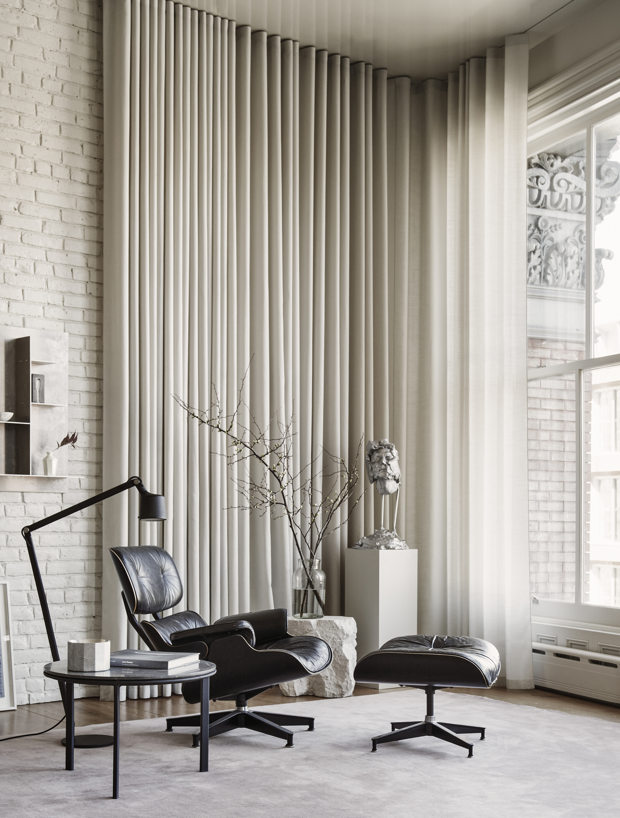eames chair and ottoman in a scandi-style room with curtains, gray rug, big windows, and a floor lamp reaching over the side table and chair