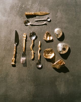 A series of golden and silver cutlery items and shiny shells sit atop a rough, black surface.