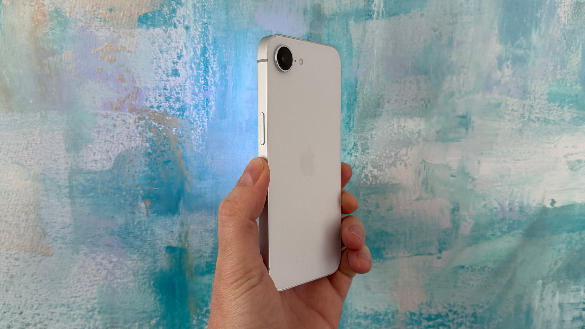 Apple iPhone 16e smartphone held in hand in front of canvas painting