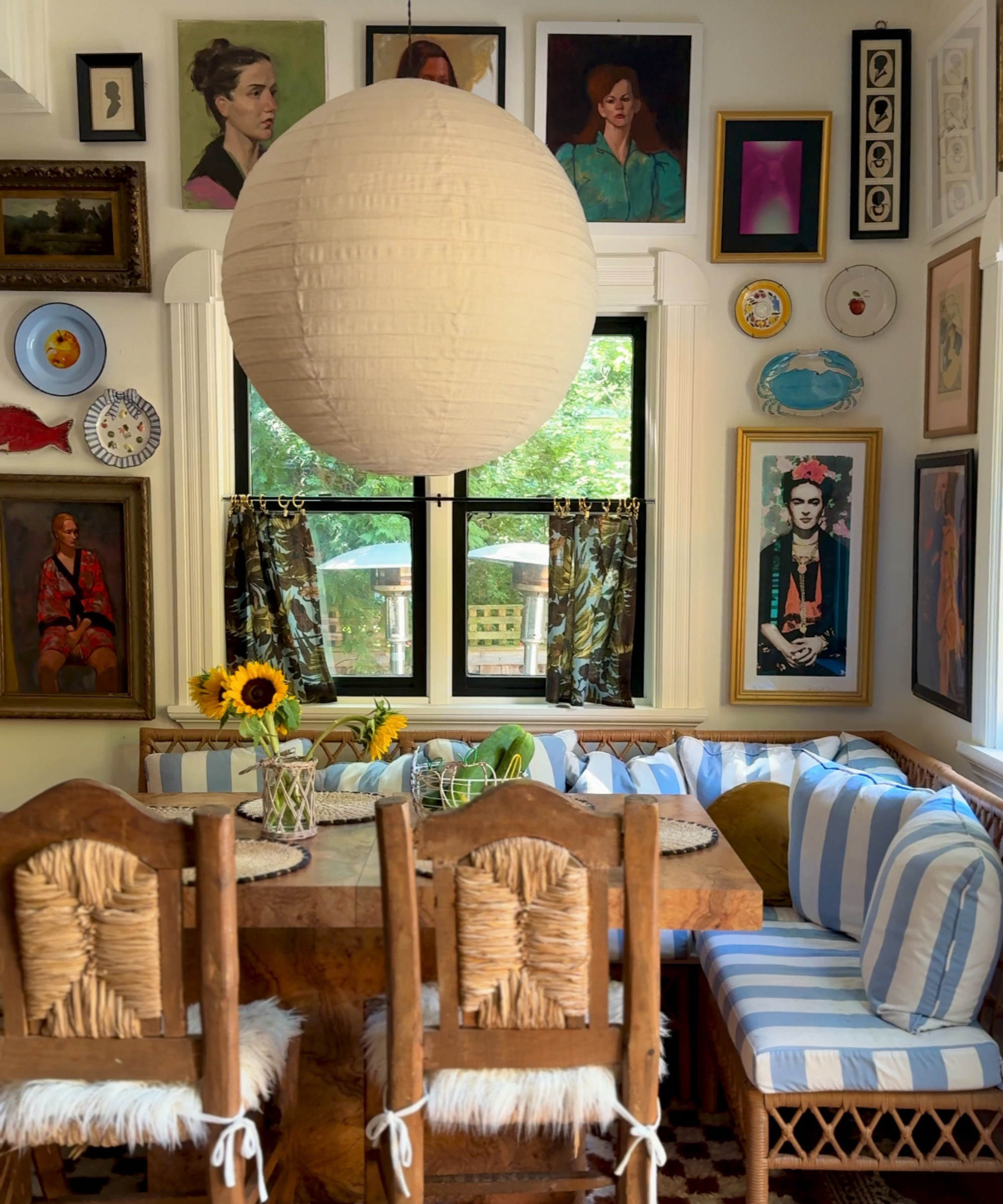 a dining nook with a corner seat with blue and white striped cushions, a square wood table, tow accent chairs, and a wall decorated with artwork. A orb paper lamp hangs over the table