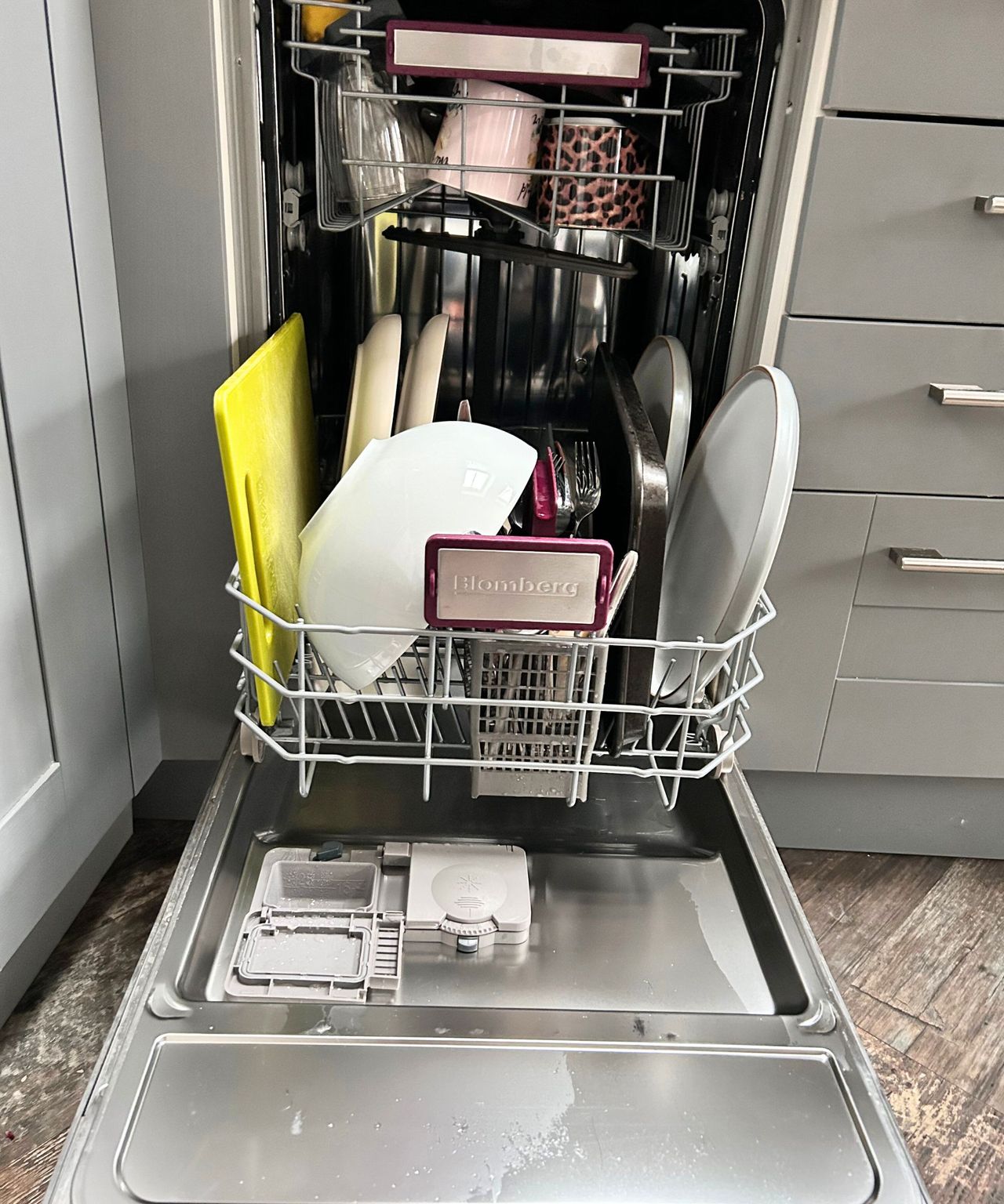 Clean dishes in a dishwasher with a green chopping board, white bowls, grey plates and cutlery in a grey kitchen