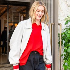 Sarah Pidgeon steps outside of a hotel wearing a red jumper with a beige jacket and black trousers. 
