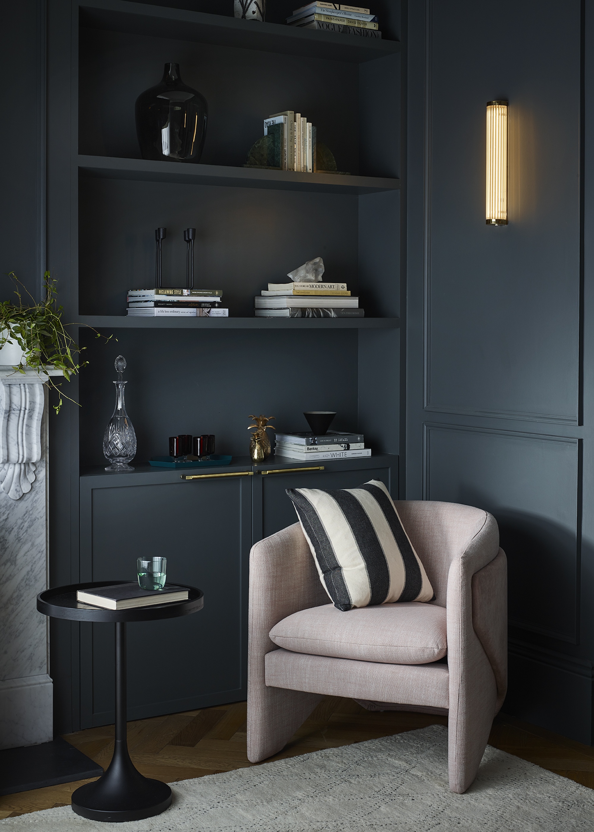 The corner of a living room painted deep dark blue with a pink chair in the corner with a cushion on top. There is also a book shelf with decor and books on top and above the chair is a wall light.