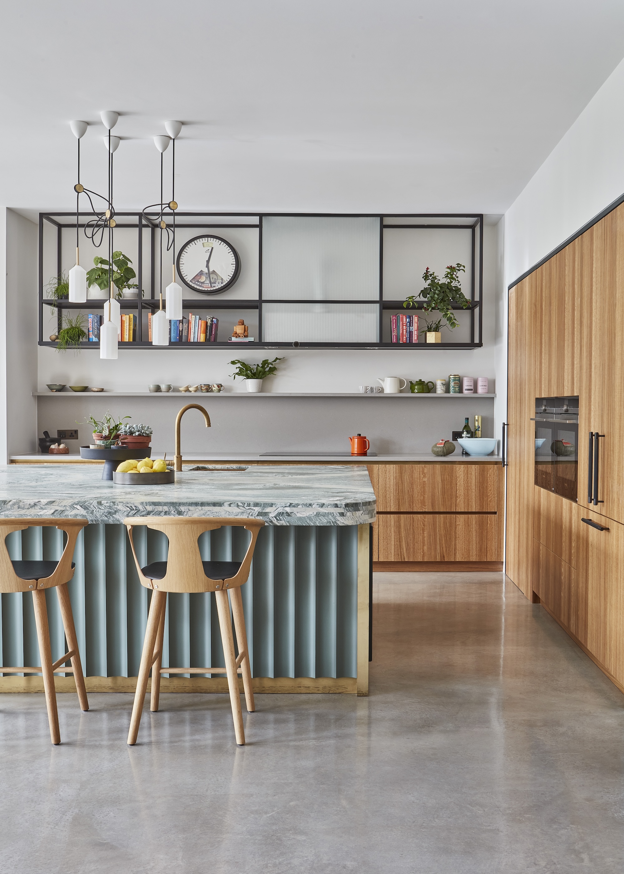 A modern kitchen with concrete floors, wooden cupboards, a stone topped island with wooden bar stools, and there is a black metal shelving unit above the hob.