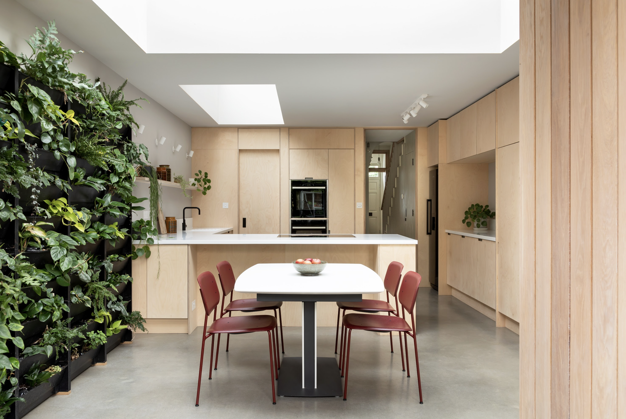 An L-shaped kitchen with a living green wall and plywood fittings, a dining table with red chairs