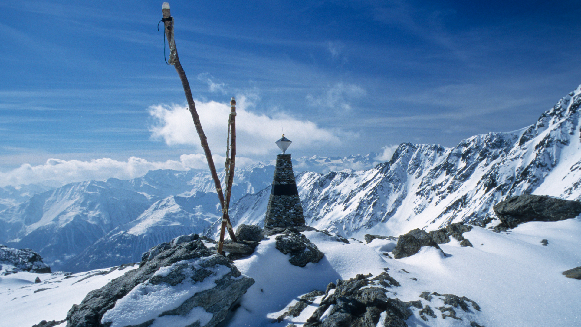 A pyramidal stone monument stands in the middle of snow-covered mountains, with a brilliant blue sky in the background