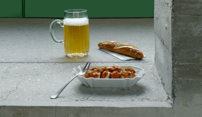 Glass of beer next to sesame seed roll and white bowl with currywurst