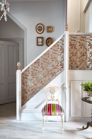A light grey hallway with light grey laminate wood-effect flooring with ornate gilded white staircase and striped velvet dining chair