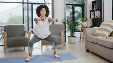 A woman performs a weighted dumbbell squat at home on a yoga mat. Her legs are bent, knees pointing outwards and her arms hold dumbbells straight out in front. Behind her we see chairs, potted plants and a couch.