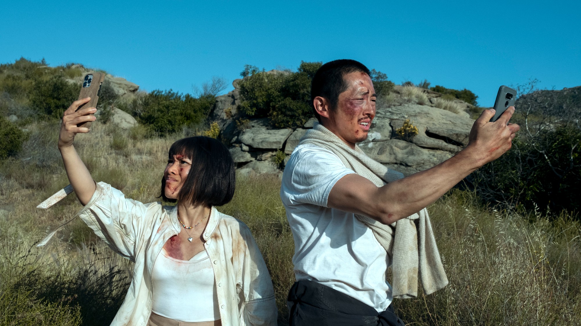 steven yeun and ali wong, both bloodied, looking into their phones out in nature