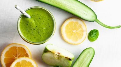 green juice next to cucumber, orange and lemon on white background