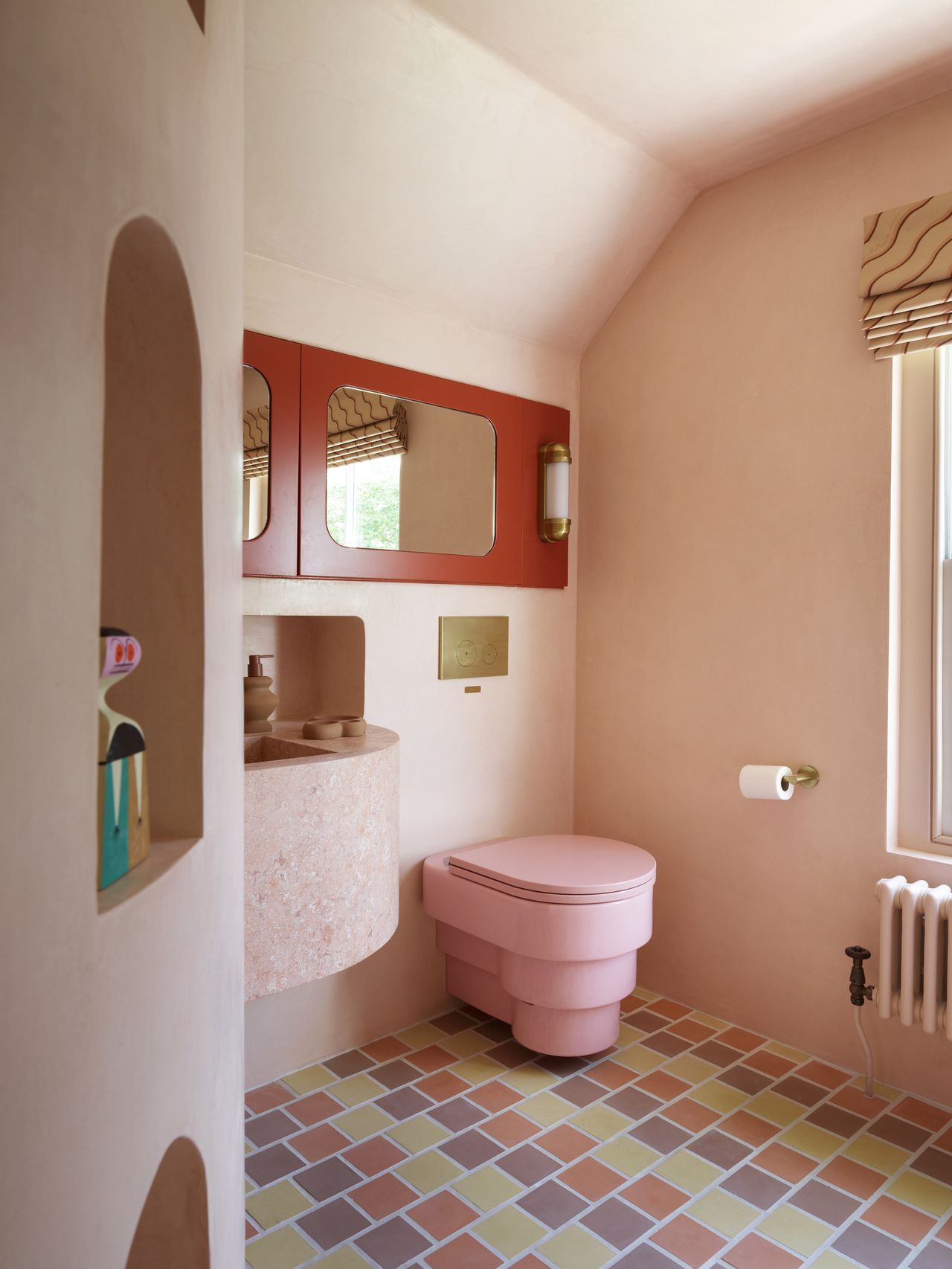 Image of a light pink bathroom with a pink toilet, a light pink terrazzo sink, and a red mirror. The floors are coral, yellow, and purple tile.