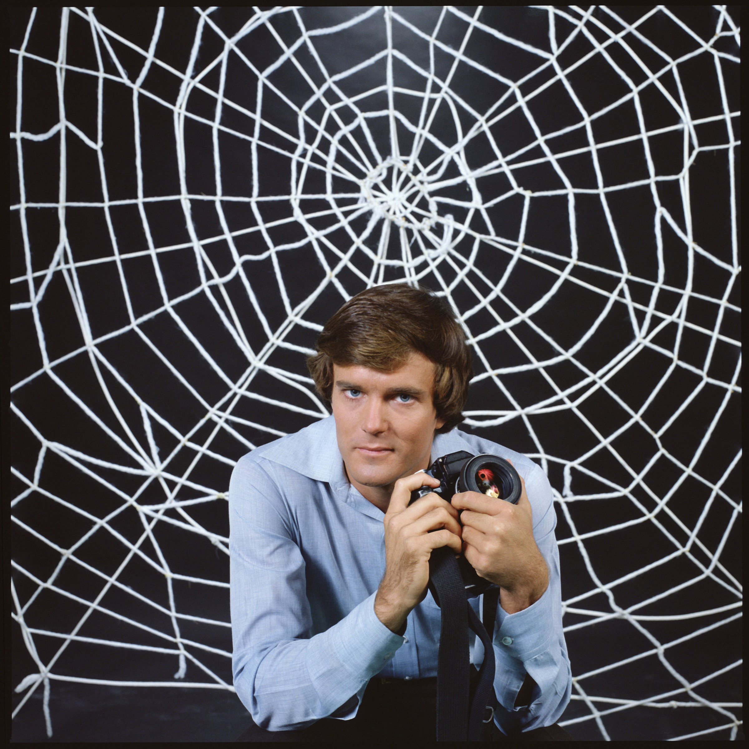 Promotional portrait of American actor Nicholas Hammond, as Peter Parker (whose secret identity is as the costumed superhero Spider-man), as he holds a camera and sits in front a large &#039;web&#039; for the CBS television series &#039;The Amazing Spider-Man,&#039; 1978.