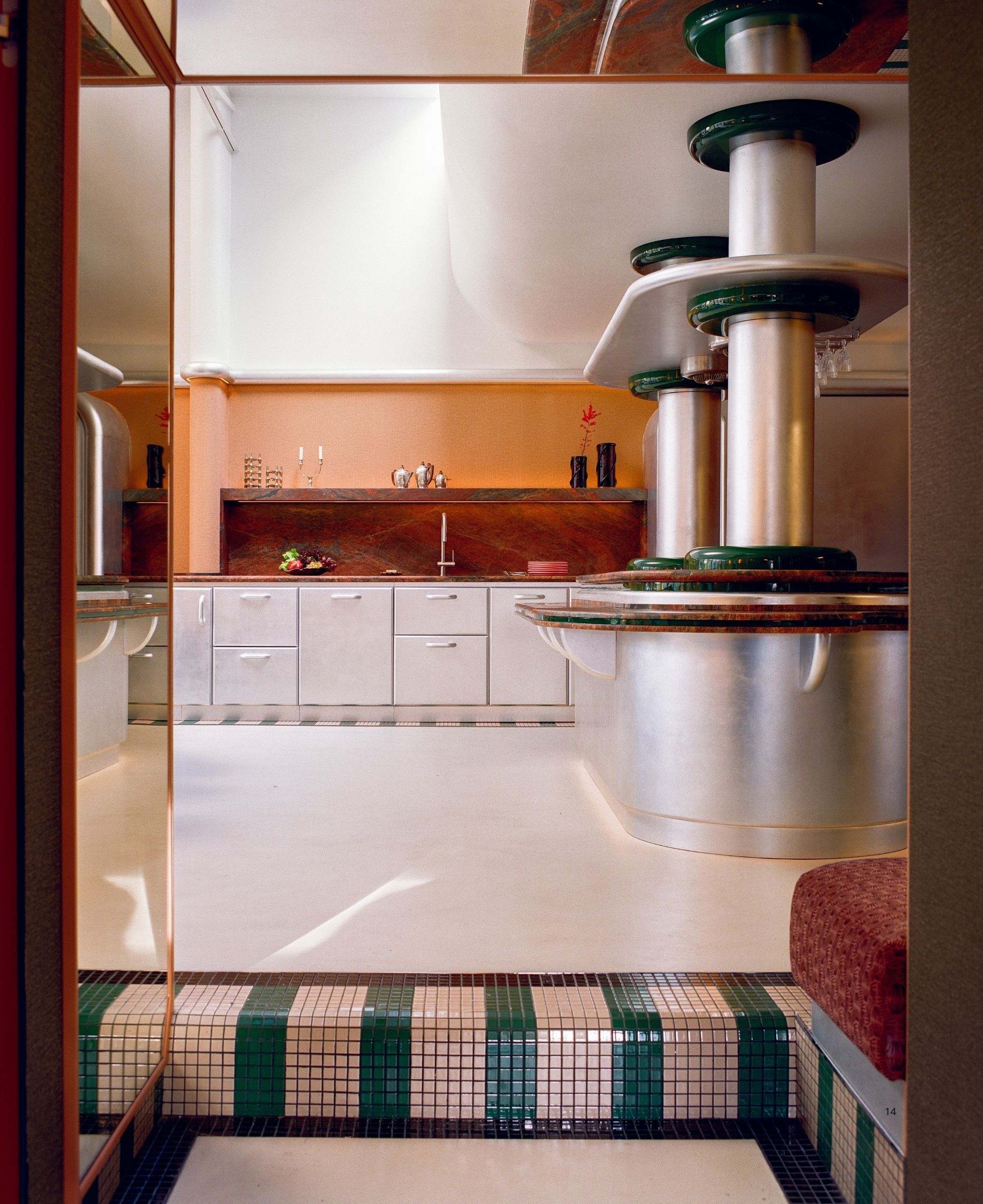 A Space Age-inspired kitchen with brushed chrome detailing throughout the room, green mosaic floor tiles, orange walls and red marble on the countertops. 