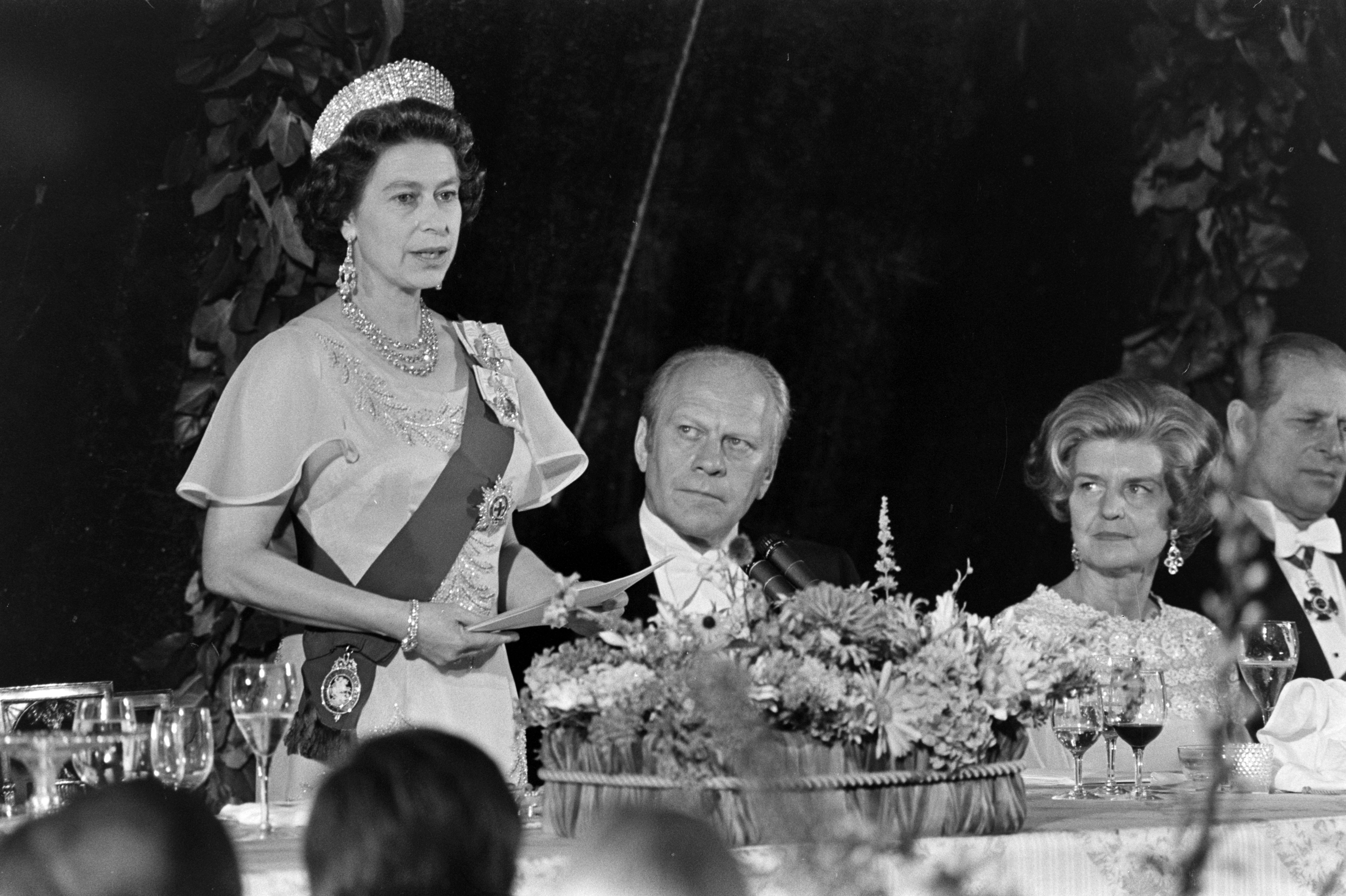 Queen Elizabeth wearing a tiara standing up and giving a speech next to Gerald and Betty Ford
