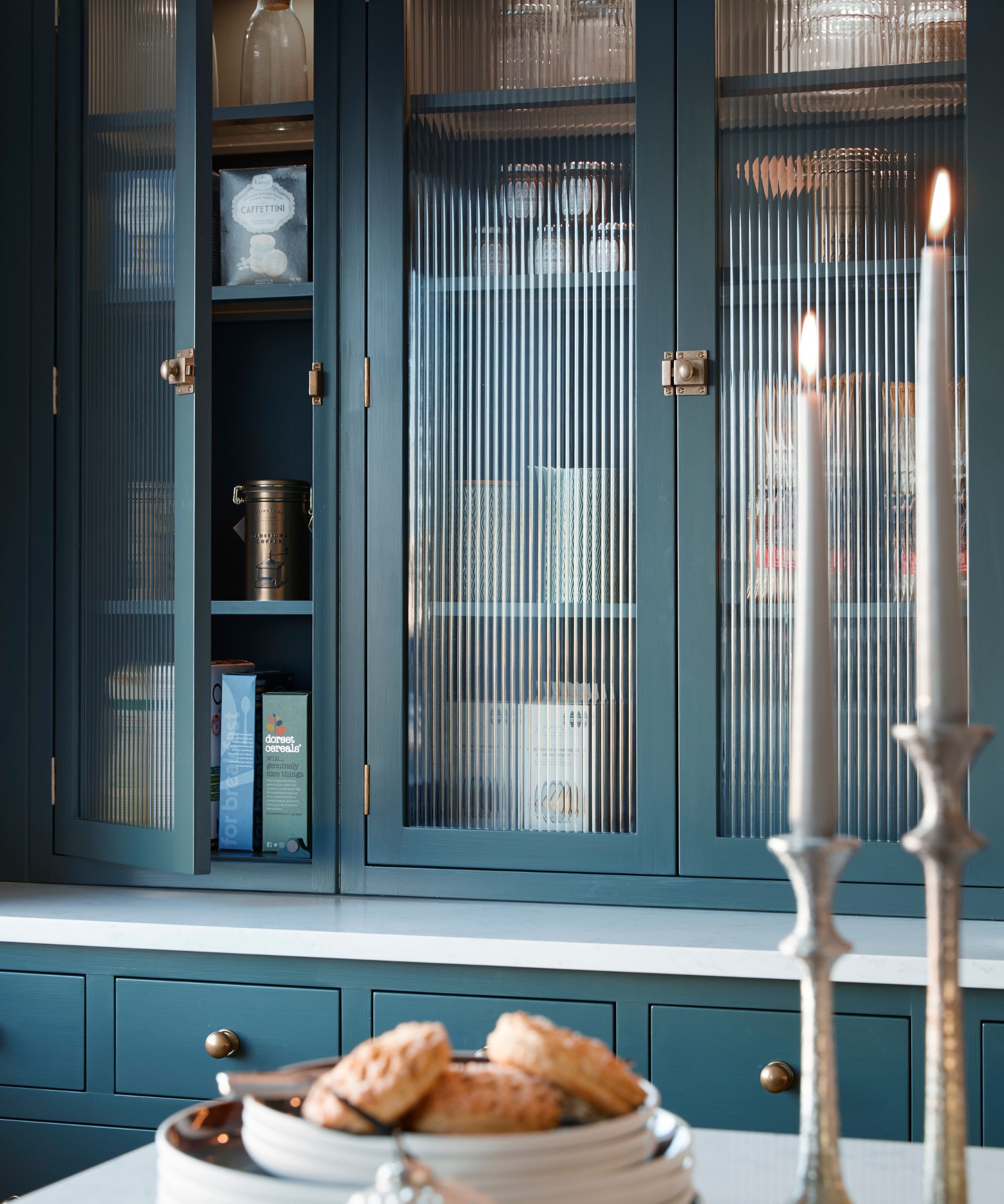 Large blue kitchen cabinets with fluted glass fronts. One door partially open to show pantry ingredients. In the foreground, blurry, a plate of small pastries and two lit candles.